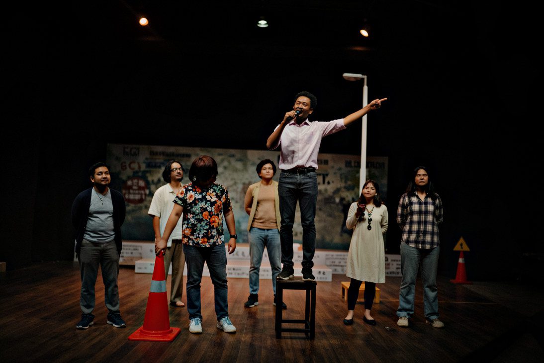 Group of performers on a theatre stage, with one actor standing on a raised platform during a dramatic scene.